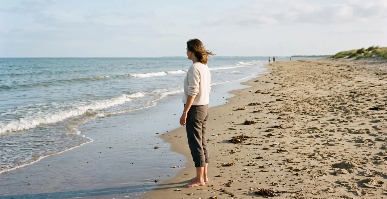 A woman stands on a calm September beach looking at the gentle sea.